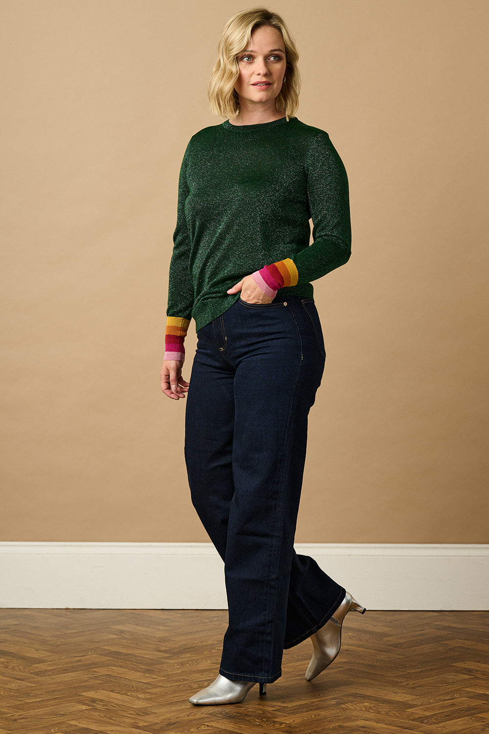 A woman wearing a green crew neck jumper with colourful striped cuffs, paired with blue jeans.
