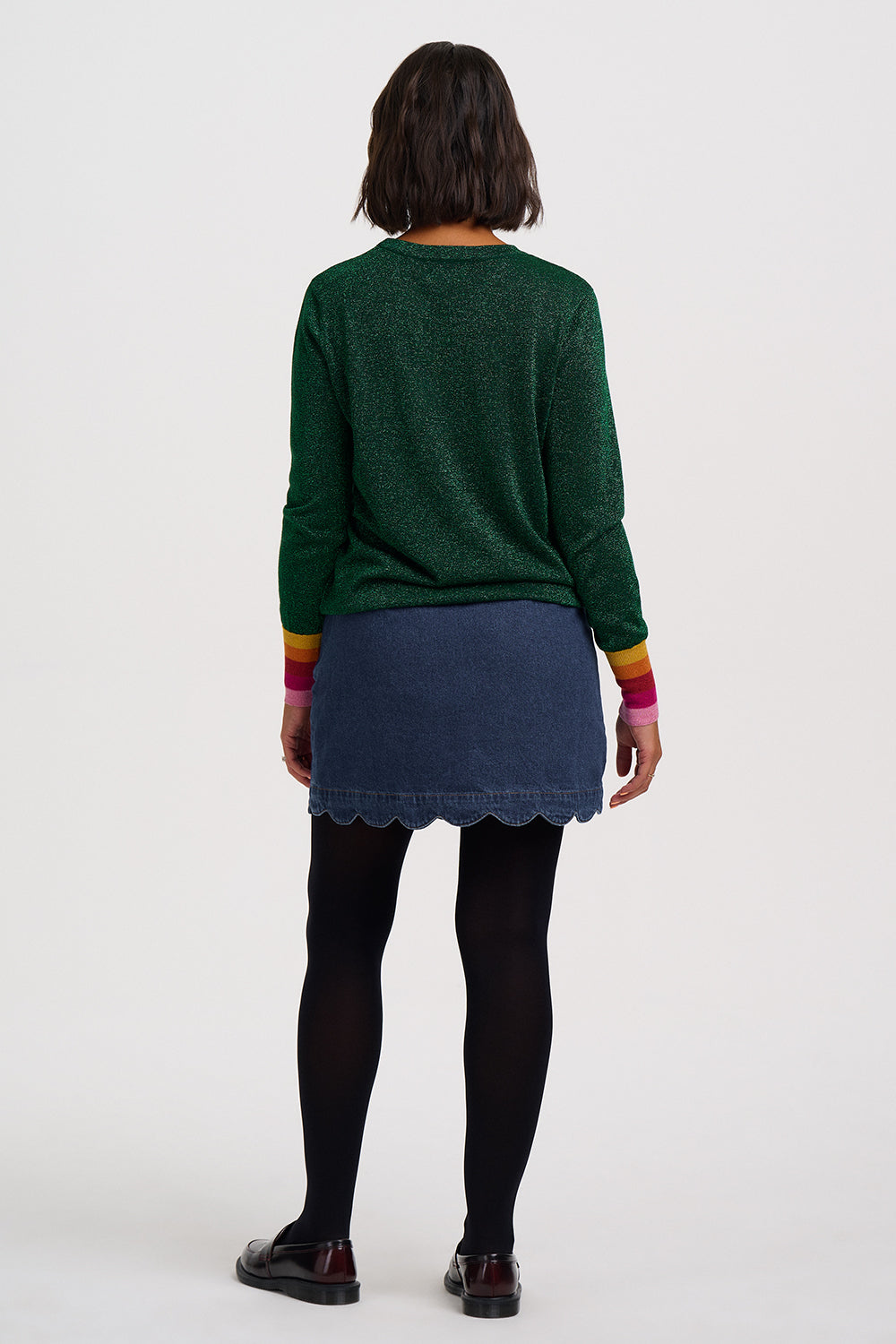 A model shown from the back wearing a dark green knit sweater with colorful striped cuffs, a dark blue denim skirt, black tights, and dark brown shoes, against a plain white studio background.