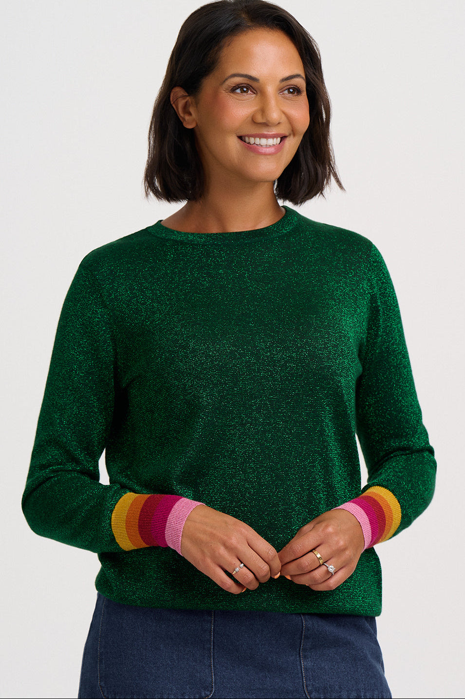 A smiling woman with short dark hair wears a dark green long-sleeve knit sweater with colorful striped cuffs in yellow, orange, pink, and green, paired with blue jeans against a light gray studio background.