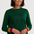 A smiling woman with short dark hair wears a dark green long-sleeve knit sweater with colorful striped cuffs in yellow, orange, pink, and green, paired with blue jeans against a light gray studio background.