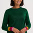 A smiling woman with short dark hair wears a dark green long-sleeve knit sweater with colorful striped cuffs in yellow, orange, pink, and green, paired with blue jeans against a light gray studio background.