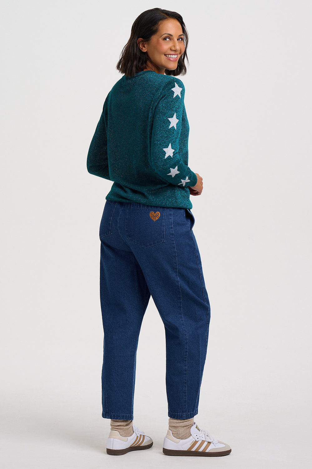 A woman modeling a dark teal knit sweater with white embroidered stars on the sleeves, paired with dark blue jeans and white sneakers, shown from a back-three-quarter angle against a light gray studio background.