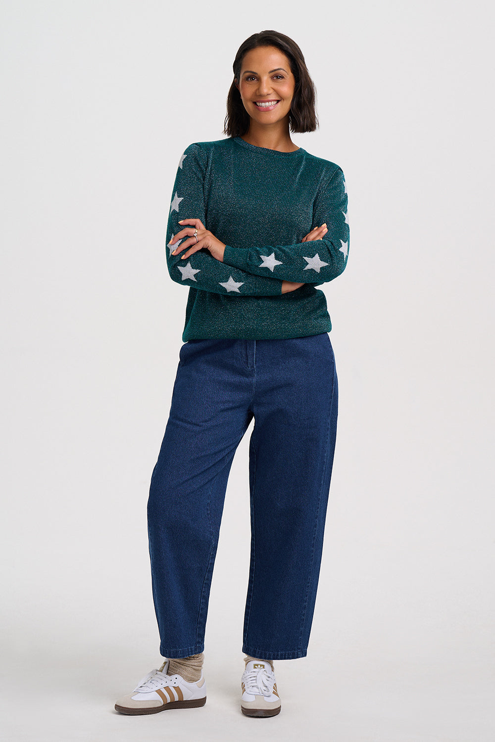 A woman modeling a dark green crewneck sweater with white star patterns on the sleeves, paired with dark blue jeans and white sneakers, against a light gray studio background.