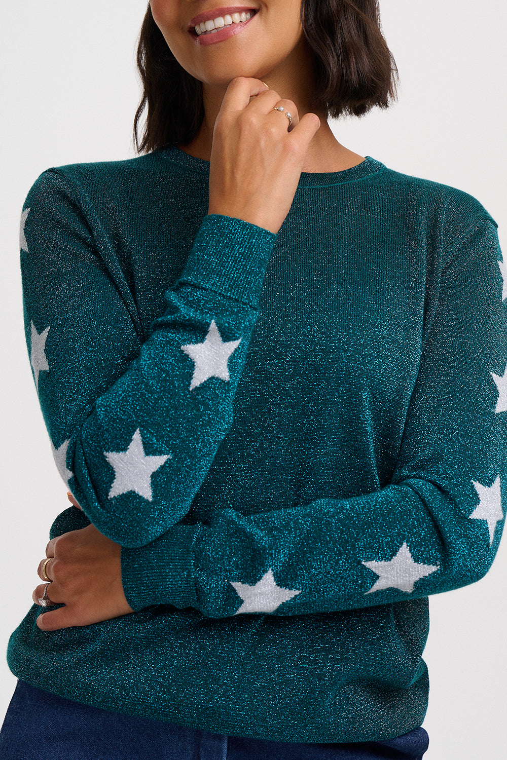 A woman wearing a dark teal long-sleeve knit sweater with white star patterns, shown from the chest up against a light gray studio background.