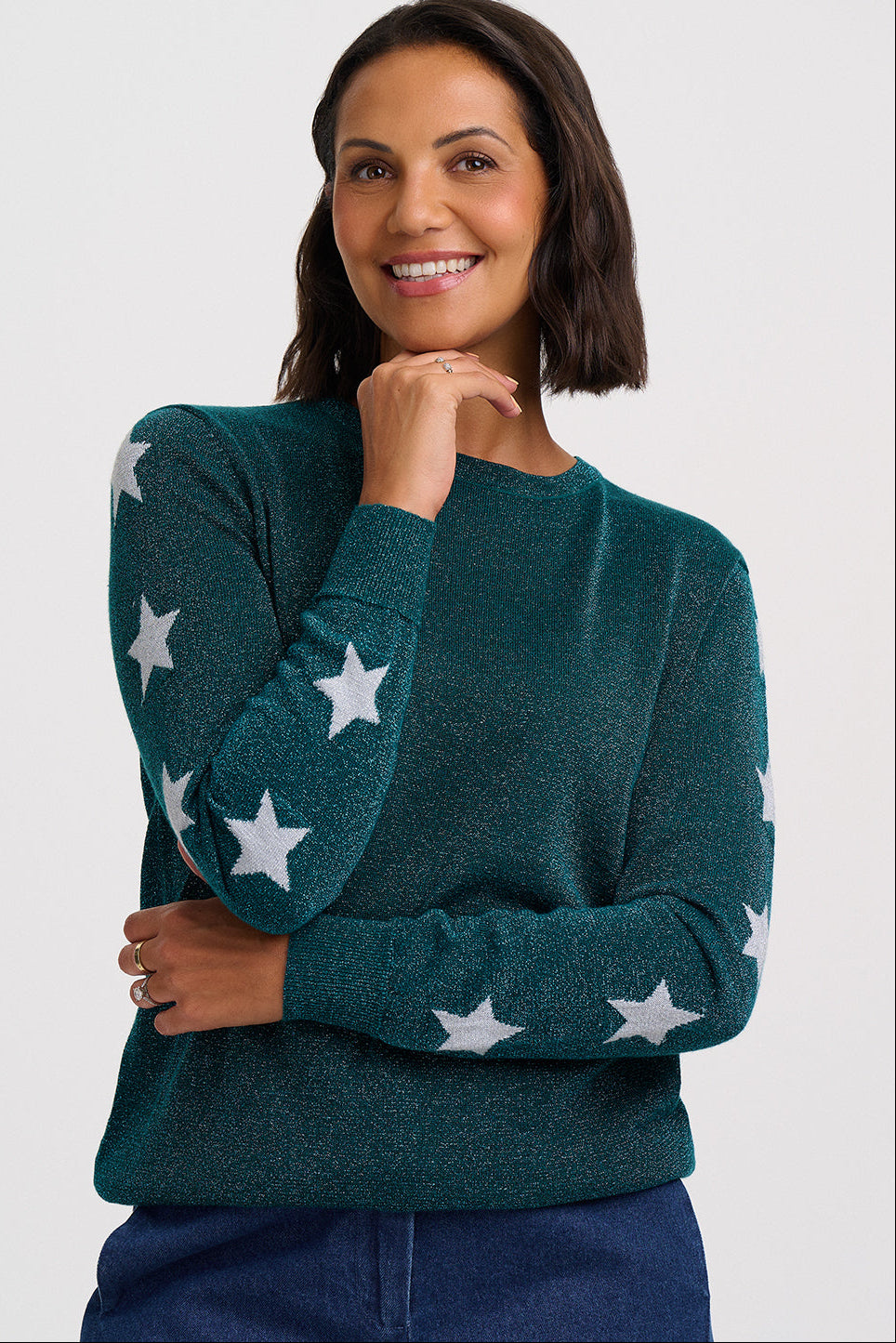 A woman wearing a dark teal knit sweater with white star patterns on the sleeves and body, paired with dark blue jeans, photographed against a light gray studio background.