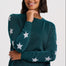 A woman wearing a dark teal knit sweater with white star patterns on the sleeves and body, paired with dark blue jeans, photographed against a light gray studio background.