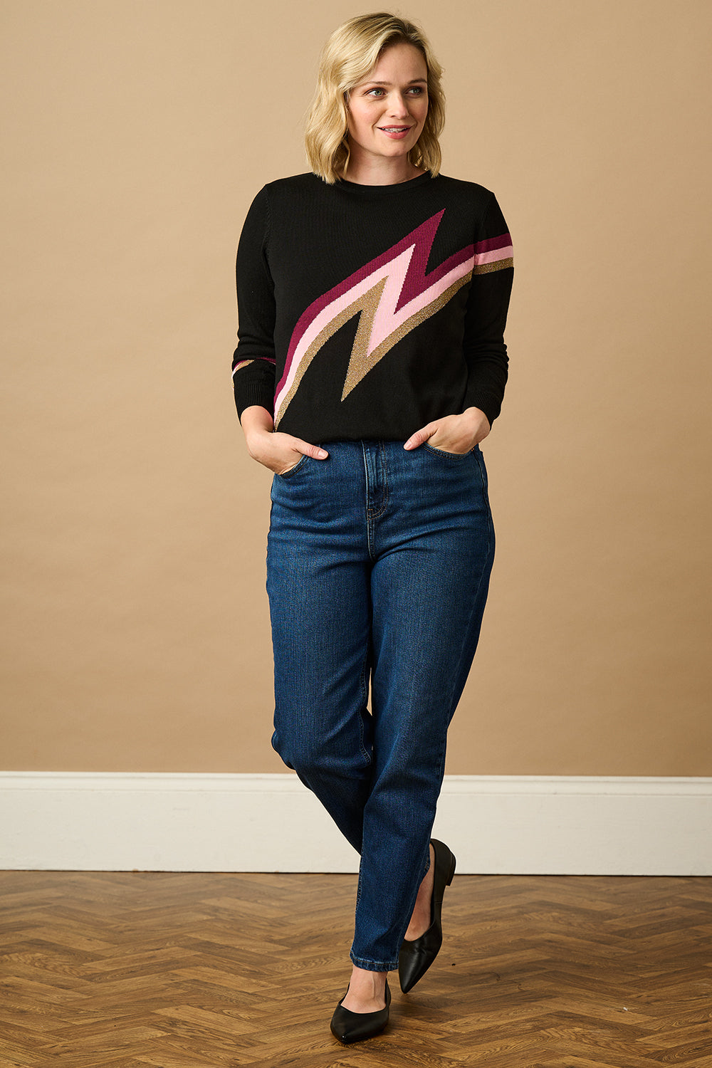 Woman wearing a black sweater with colorful stripes and blue jeans against a beige background