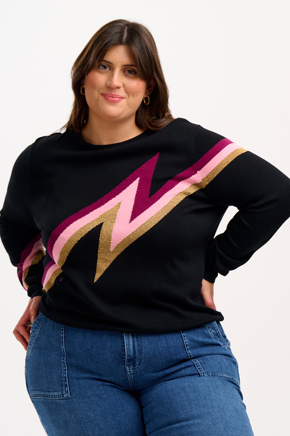 A woman wearing a long sleeve, mid-hip length black jumper with a geometric lightning bolt pattern in pink and gold across the torso and arms.