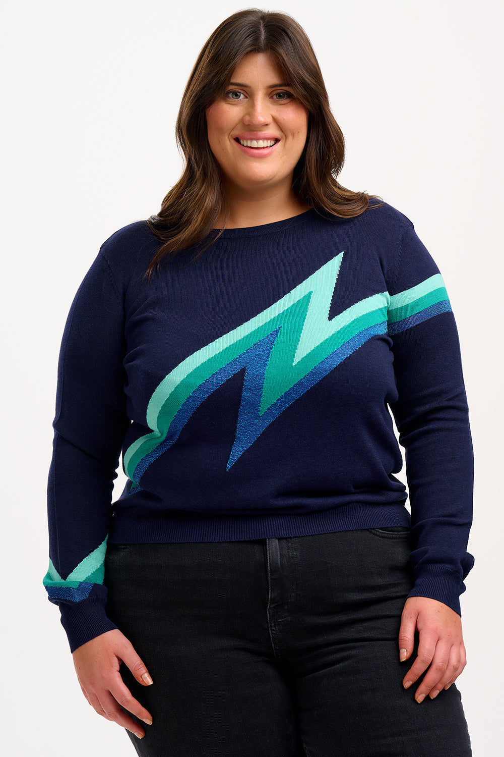 Woman wearing a navy sweater with a green lightning bolt design and blue jeans on a white background