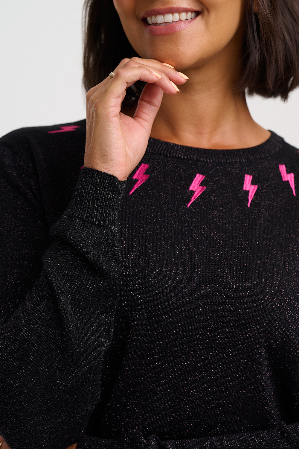 A close-up of a woman wearing a black knit sweater with bright pink lightning bolt patterns across the chest, against a light gray background.