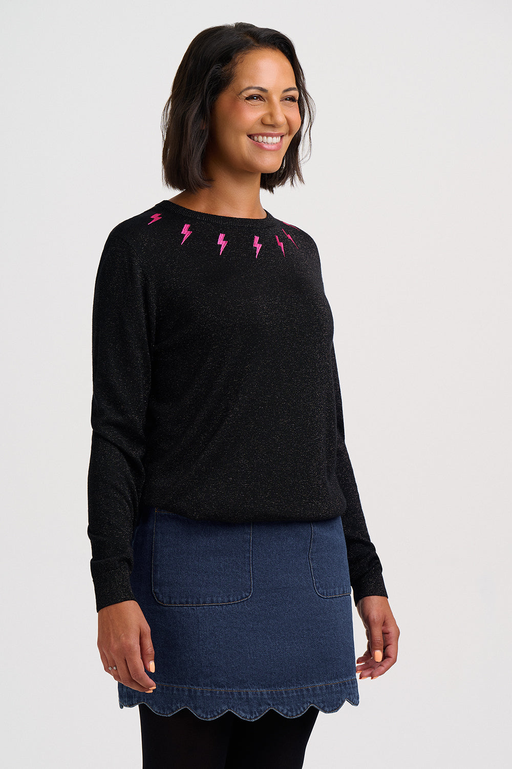 A woman wearing a black long-sleeve knit sweater with pink lightning-bolt embroidery around the neckline, paired with a blue denim skirt, against a light gray studio background.