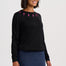 A woman wearing a black long-sleeve knit sweater with pink lightning-bolt embroidery around the neckline, paired with a blue denim skirt, against a light gray studio background.