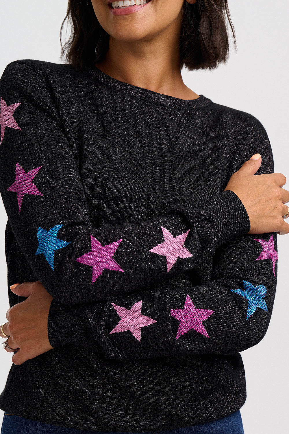A close-up of a woman wearing a black long-sleeve knit sweater with glittery pink and blue star patterns on the sleeves, against a light gray background.