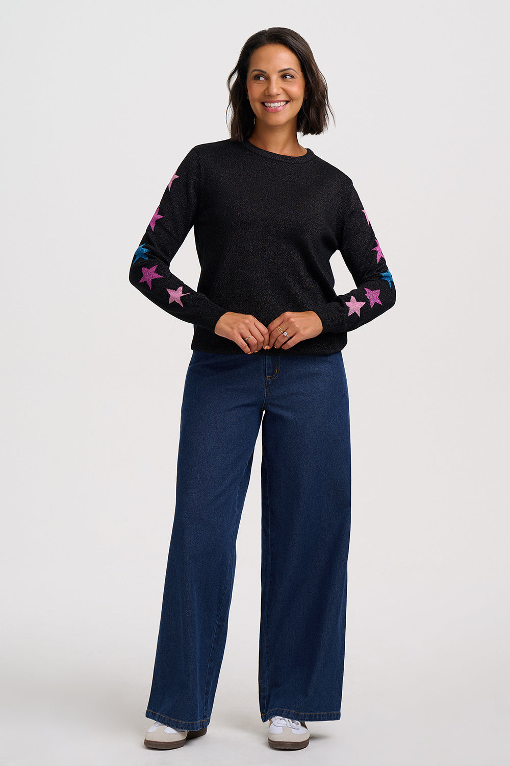 A woman modeling a black long-sleeve knit sweater with pink and blue star patterns on the sleeves, paired with dark blue wide-leg jeans and white sneakers, against a light gray studio background.