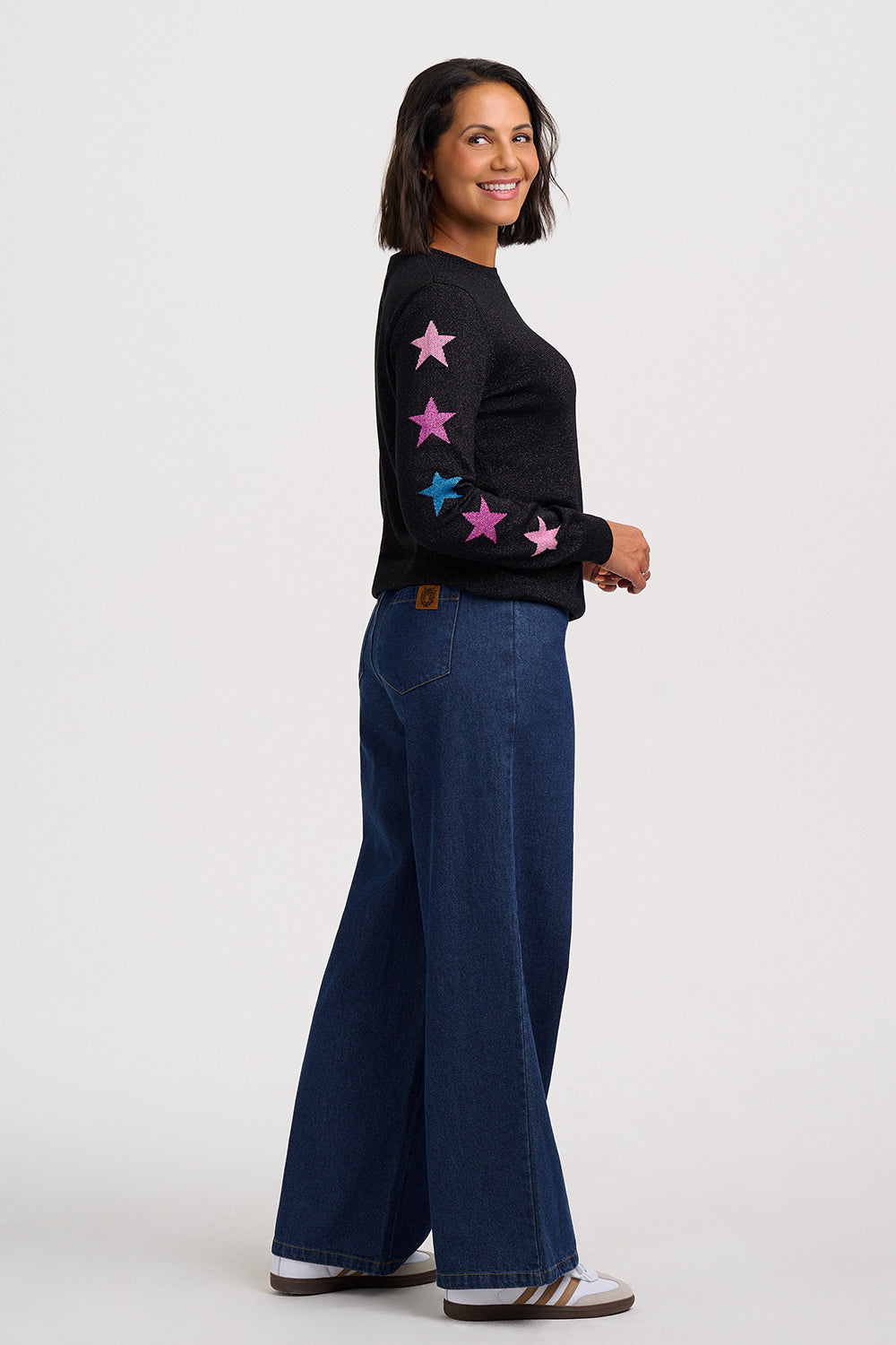 A woman modeling a black knit sweater with pink and blue star patches on the sleeves, paired with dark blue wide-leg jeans and white sneakers, against a light gray studio background.