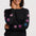 A woman wearing a black crewneck sweater with pink and blue star patterns on the sleeves, paired with blue jeans, against a light gray studio background.