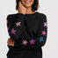 A woman wearing a black crewneck sweater with pink and blue star patterns on the sleeves, paired with blue jeans, against a light gray studio background.