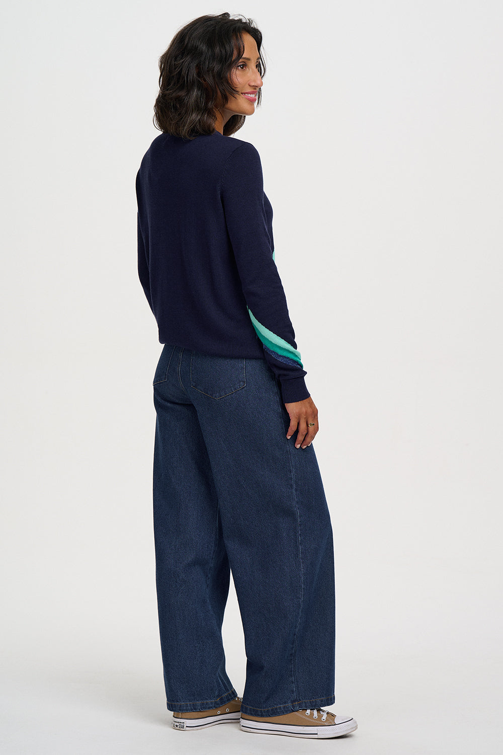 A woman shown from the back wearing a navy blue long-sleeve knit sweater with a teal and light blue striped accent on the sleeve, paired with wide-leg dark blue jeans and brown sneakers, against a plain white studio background.