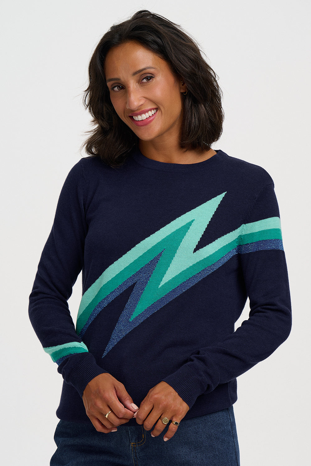 A woman wearing a navy blue crewneck sweater with a large diagonal lightning bolt pattern in shades of green and teal across the chest, paired with dark blue jeans, against a light gray studio background.