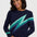 A woman wearing a navy blue crewneck sweater with a large diagonal lightning bolt pattern in shades of green and teal across the chest, paired with dark blue jeans, against a light gray studio background.