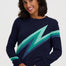 A woman wearing a navy blue crewneck sweater with a large diagonal lightning bolt pattern in shades of green and teal across the chest, paired with dark blue jeans, against a light gray studio background.