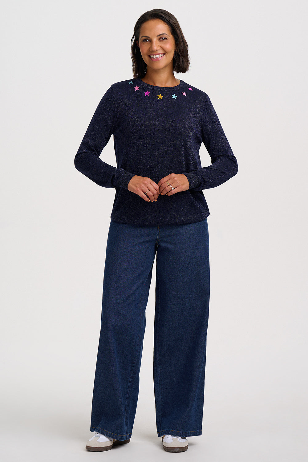 A woman modeling a dark navy blue knit sweater with colorful embroidered stars at the neckline, paired with dark blue wide-leg pants, against a plain light gray studio background.