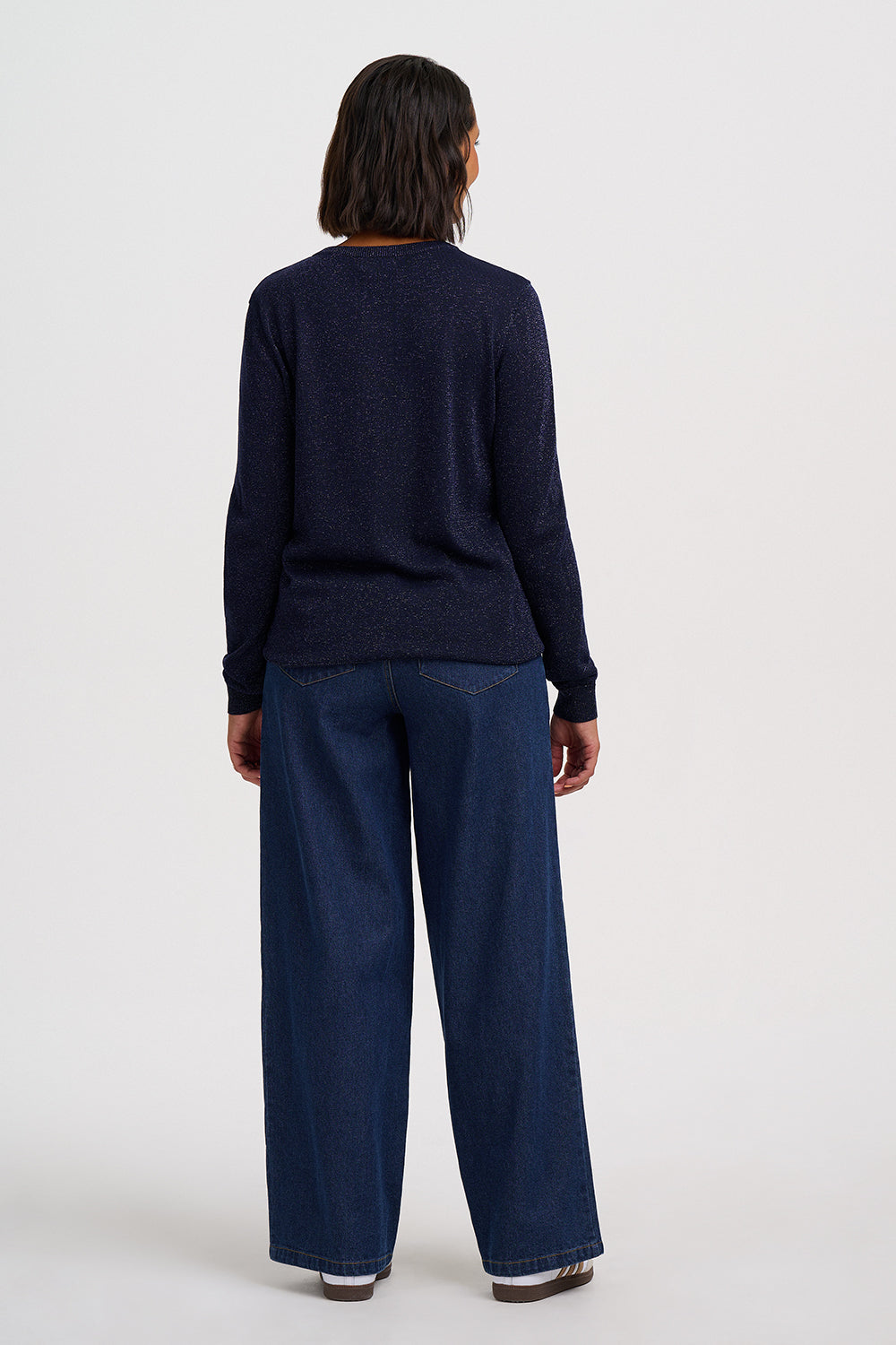 A model shown from the back wearing a dark navy blue knit sweater and wide-leg dark blue jeans, standing against a plain light gray studio background.