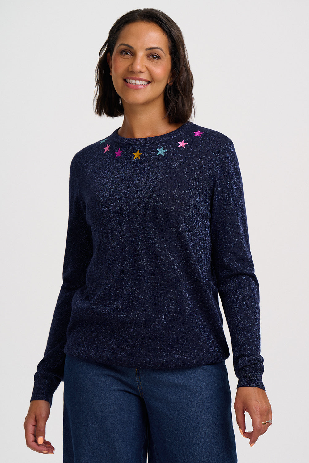 A woman wearing a dark navy blue knit sweater with small embroidered stars in pink, yellow, and blue around the neckline, paired with dark blue jeans, against a light gray studio background.