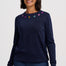 A woman wearing a dark navy blue knit sweater with small embroidered stars in pink, yellow, and blue around the neckline, paired with dark blue jeans, against a light gray studio background.