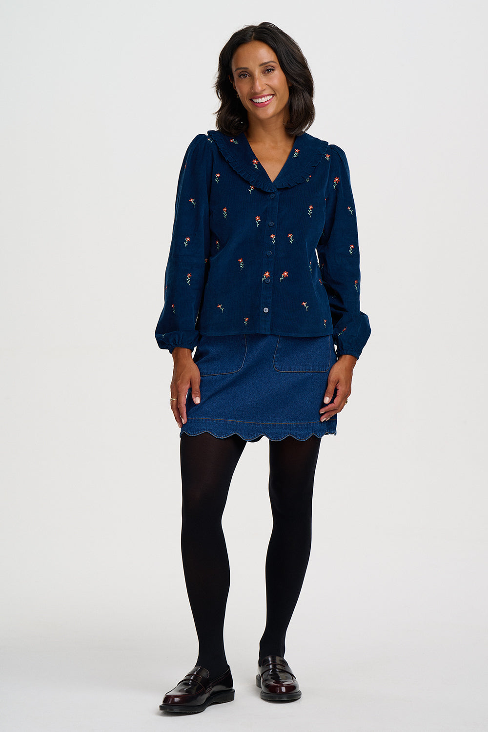 A woman modeling a navy blue velvet long-sleeve top and matching mini skirt set with embroidered details, paired with black tights and dark brown leather shoes, against a light gray studio background.