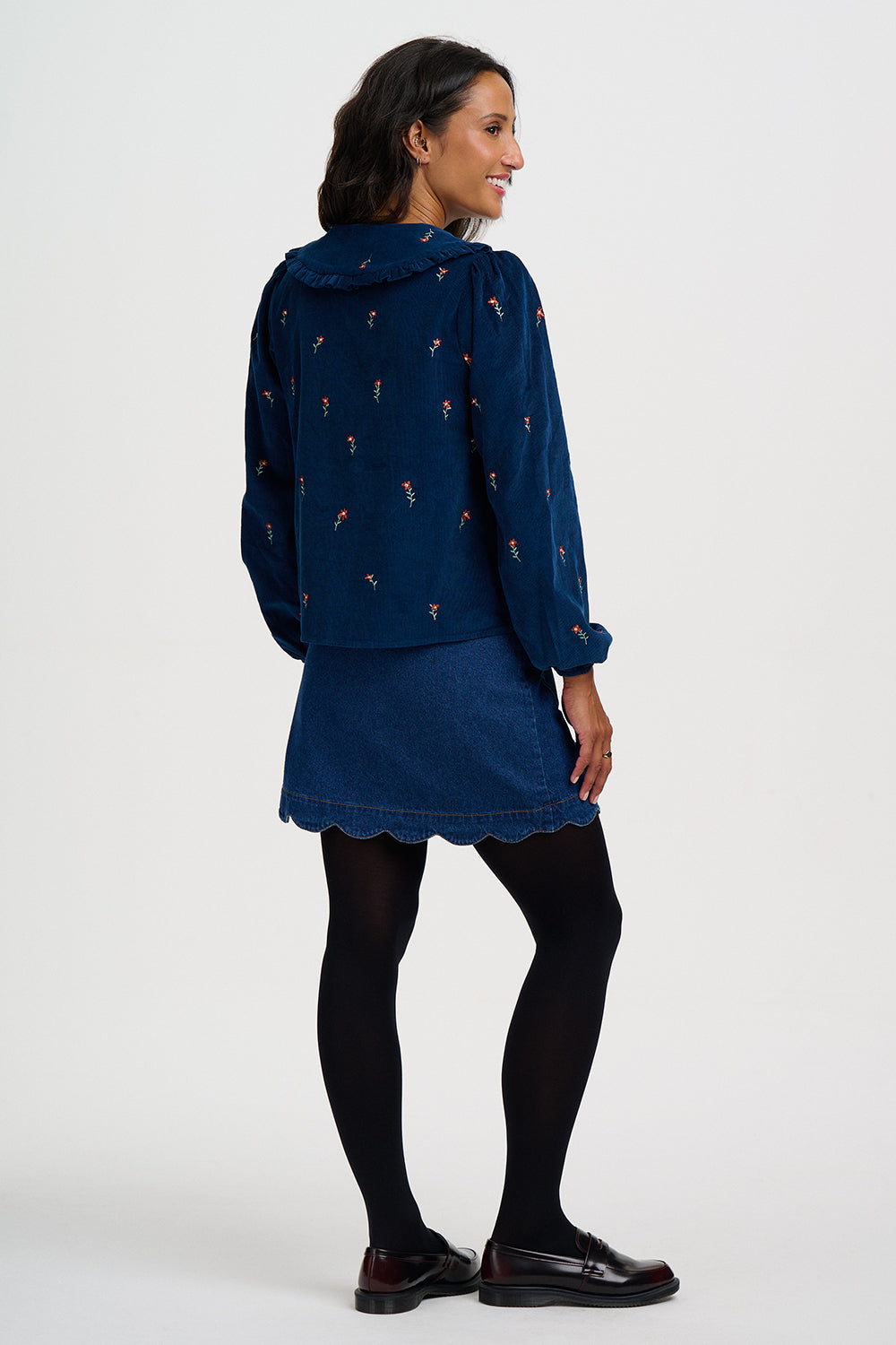 A woman is shown from the back wearing a navy blue hooded jacket with a small floral pattern, a matching denim-look mini skirt, black tights, and dark loafers, against a plain light gray studio background.