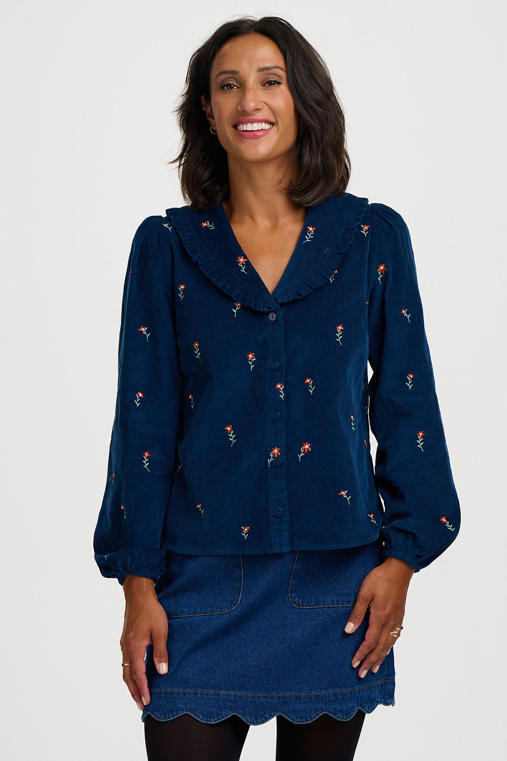 A woman modeling a navy blue long-sleeve button-up shirt with small embroidered floral details, paired with a matching denim mini skirt, against a light gray studio background.