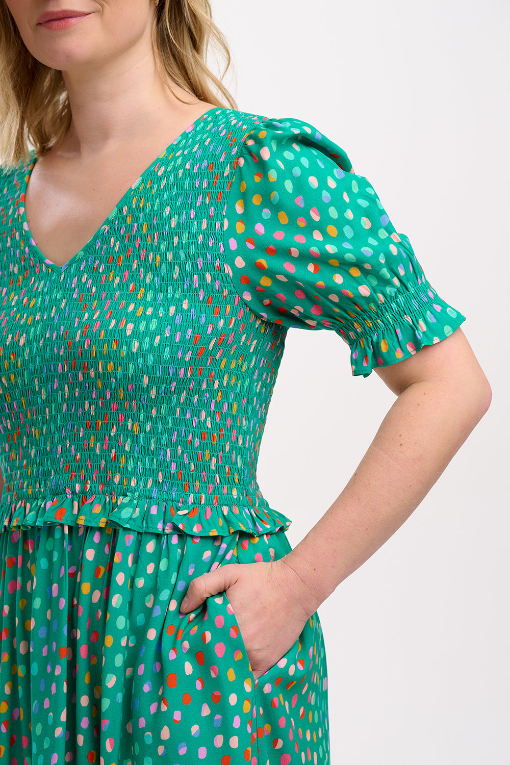A woman standing wearing a green midi dress with a shirred bodice, short shirred sleeves, and a waist frill, printed with a pattern of rainbow spots.