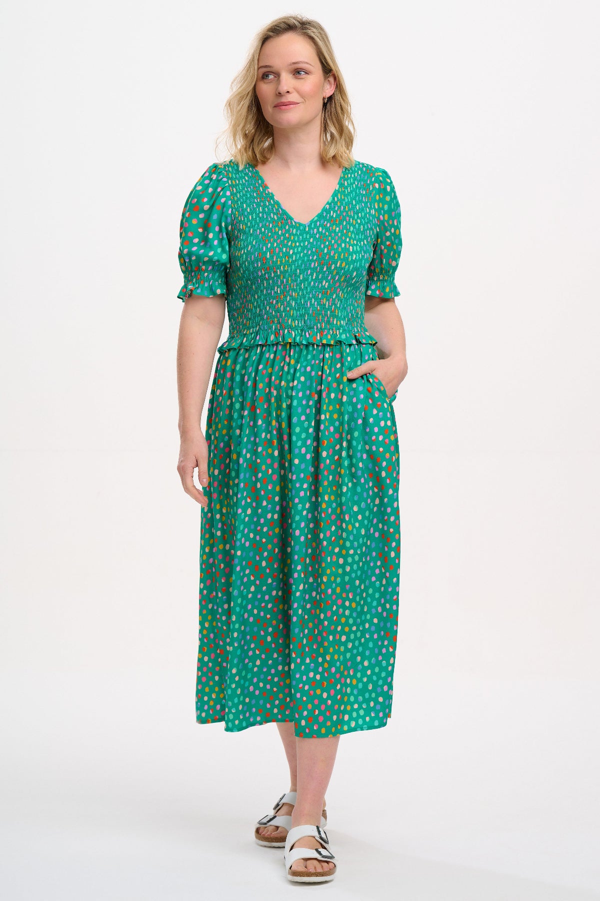 A woman standing wearing a green midi dress with a shirred bodice, short shirred sleeves, and a waist frill, printed with a pattern of rainbow spots.