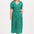 A woman standing wearing a green midi dress with a shirred bodice, short shirred sleeves, and a waist frill, printed with a pattern of rainbow spots.