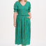 A woman standing wearing a green midi dress with a shirred bodice, short shirred sleeves, and a waist frill, printed with a pattern of rainbow spots.
