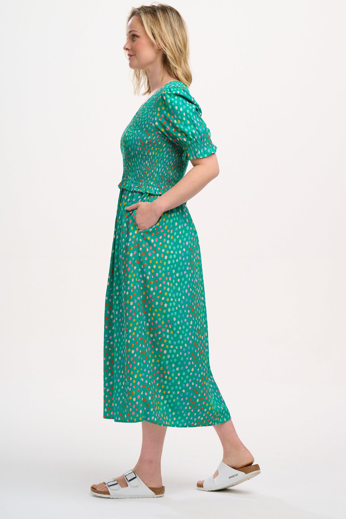 A woman standing wearing a green midi dress with a shirred bodice, short shirred sleeves, and a waist frill, printed with a pattern of rainbow spots.