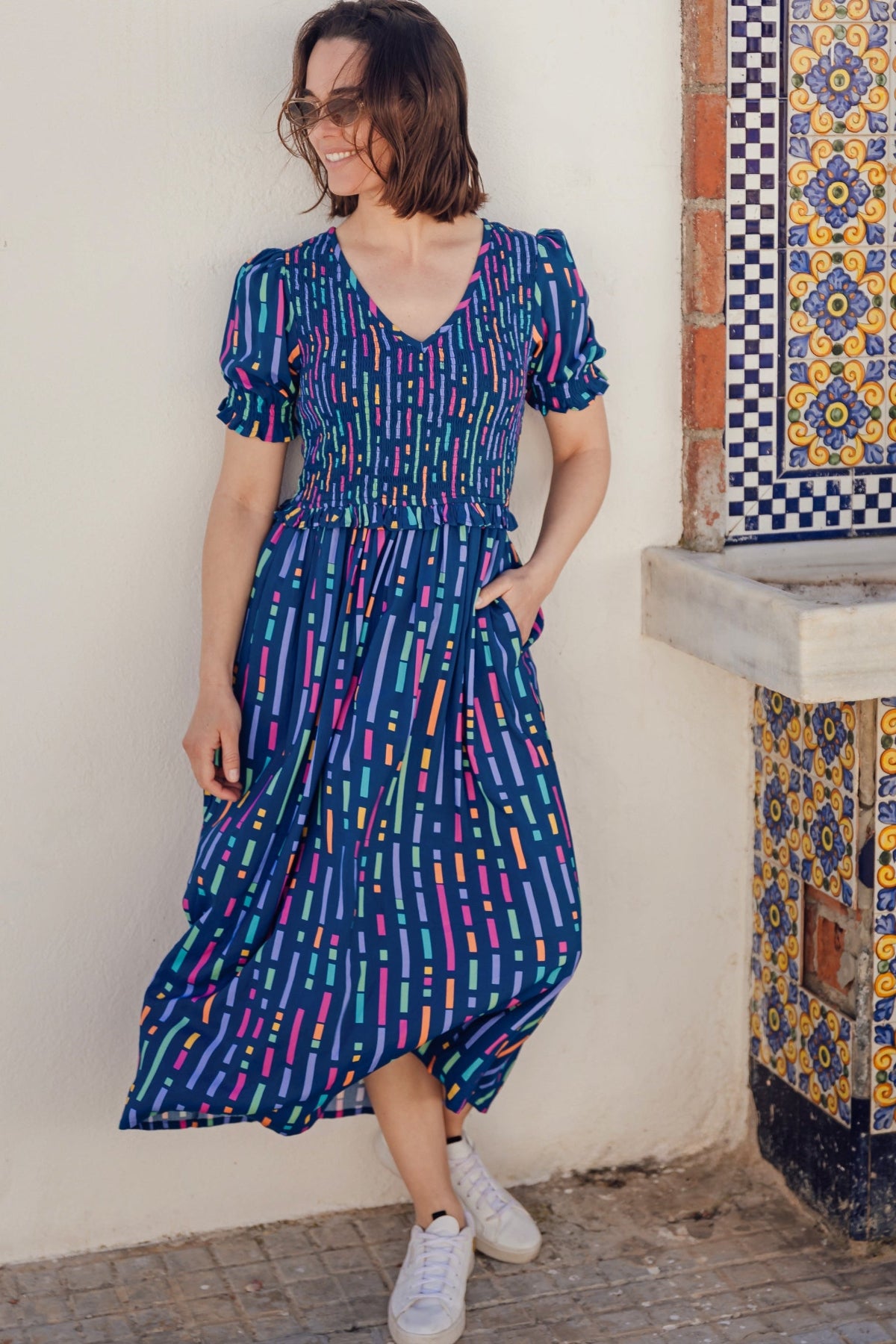A model wearing a midi length dress with a shirred bodice, waist frill, and sleeves, featuring a geometric pattern in blue and other colors on a teal background.