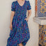 A model wearing a midi length dress with a shirred bodice, waist frill, and sleeves, featuring a geometric pattern in blue and other colors on a teal background.