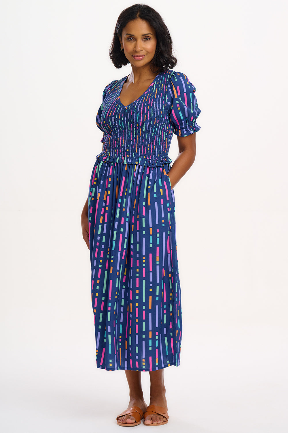 A model wearing a midi length dress with a shirred bodice, short shirred sleeves, and a V-neck. The dress has a geometric pattern with horizontal lines in blue, purple, pink, and orange.