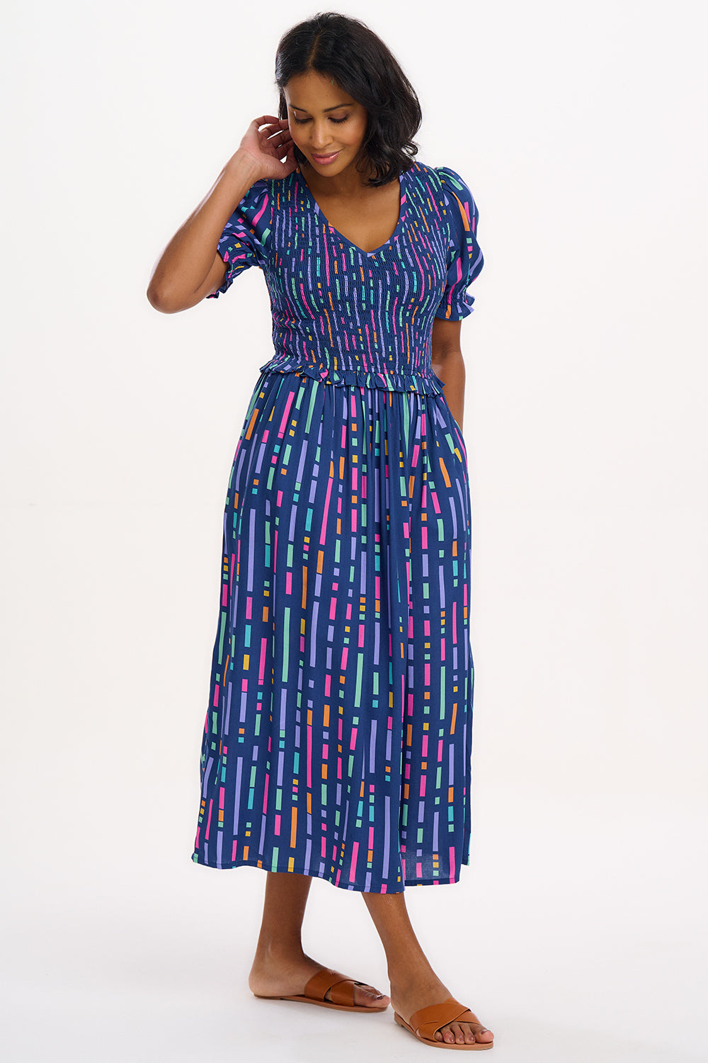 A model wearing a midi length dress with a shirred bodice, short shirred sleeves, and a V-neck. The dress has a geometric pattern with horizontal lines in blue, purple, pink, and orange.