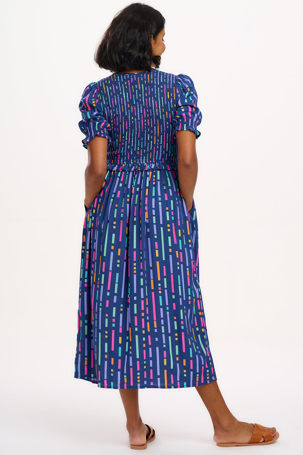 A model wearing a midi length dress with a shirred bodice, short shirred sleeves, and a V-neck. The dress has a geometric pattern with horizontal lines in blue, purple, pink, and orange.