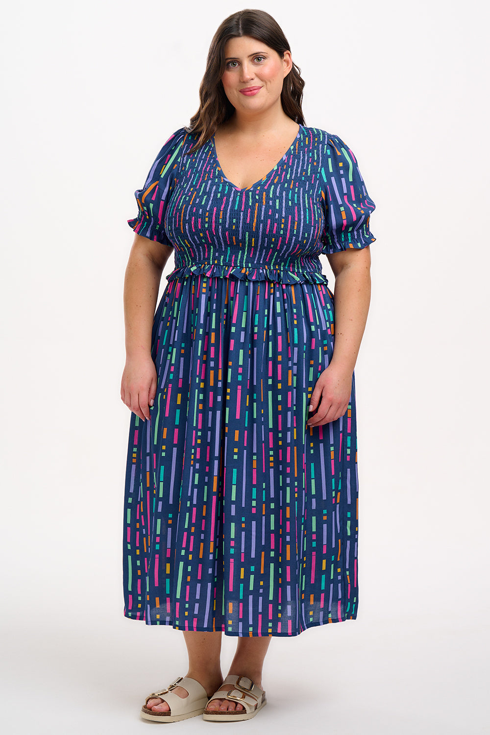 A model wearing a midi length dress with a shirred bodice, short shirred sleeves, and a V-neck. The dress has a geometric pattern with horizontal lines in blue, purple, pink, and orange.