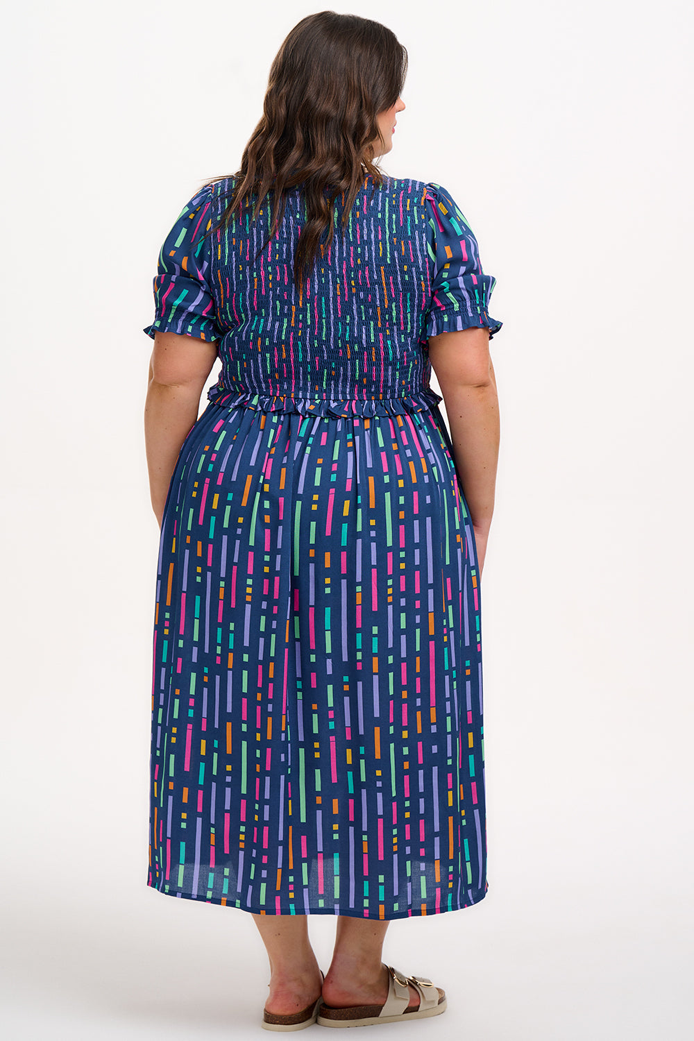 A model wearing a midi length dress with a shirred bodice, short shirred sleeves, and a V-neck. The dress has a geometric pattern with horizontal lines in blue, purple, pink, and orange.