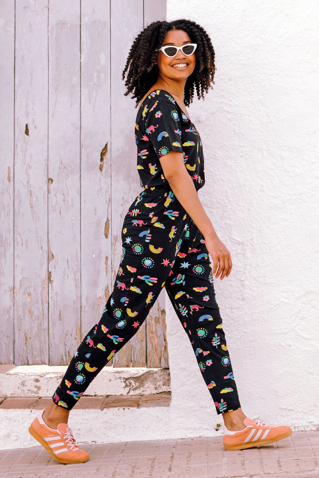 A woman wearing a black cotton jersey jumpsuit with a floral pattern, featuring short sleeves, a scoop neckline, and an elasticated waistband. She is paired with casual shoes and accessorized with sunglasses and a bracelet.