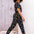 A woman wearing a black cotton jersey jumpsuit with a floral pattern, featuring short sleeves, a scoop neckline, and an elasticated waistband. She is paired with casual shoes and accessorized with sunglasses and a bracelet.