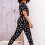 A woman wearing a black cotton jersey jumpsuit with a floral pattern, featuring short sleeves, a scoop neckline, and an elasticated waistband. She is paired with casual shoes and accessorized with sunglasses and a bracelet.