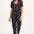 A female model wearing a black cotton jersey jumpsuit with a floral and animal print, featuring short sleeves, a scoop neckline, and elasticated waistband.