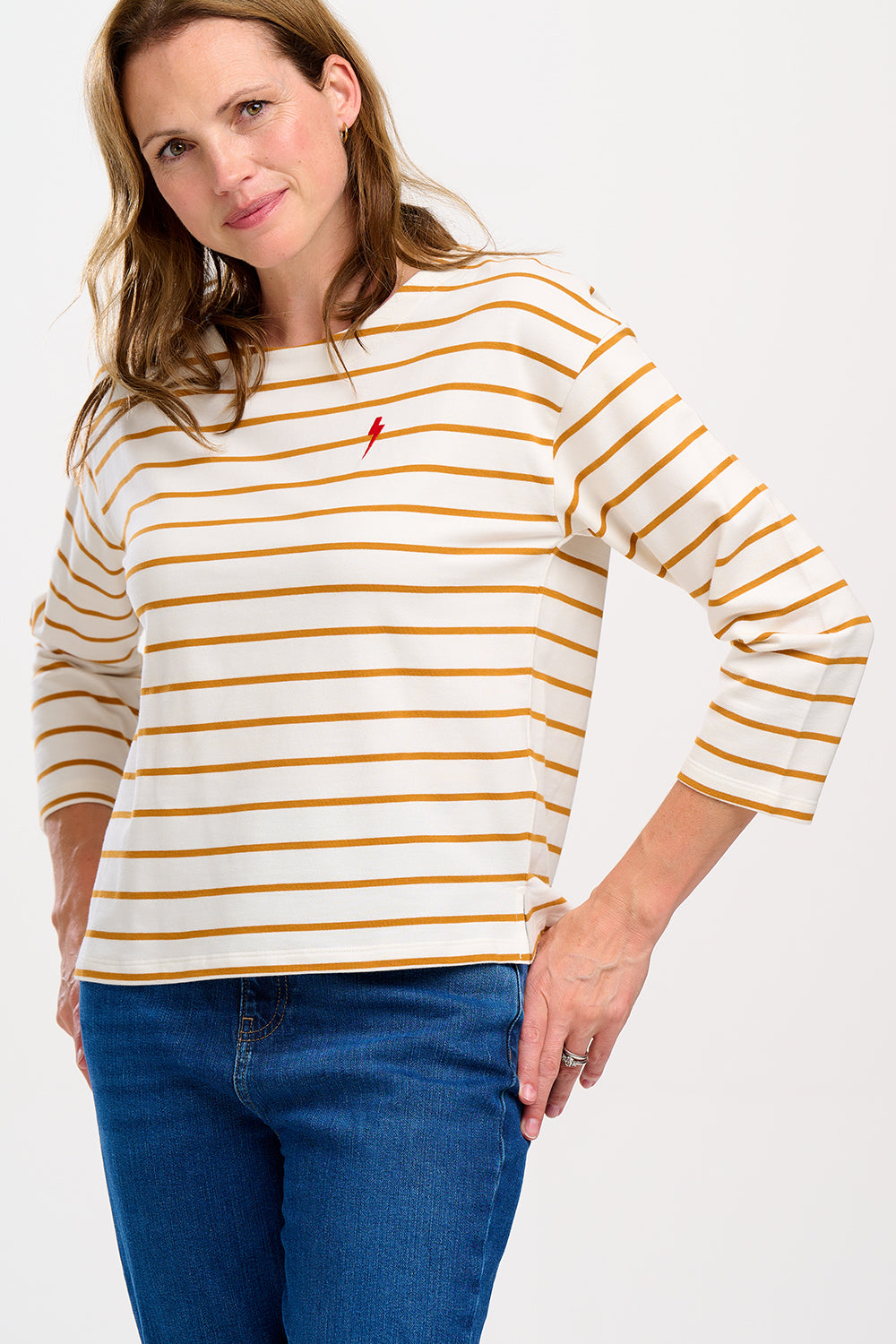 A female model wearing an off-white and tan striped Billie Breton top with a pink embroidered lightning flash on the chest, paired with blue jeans.