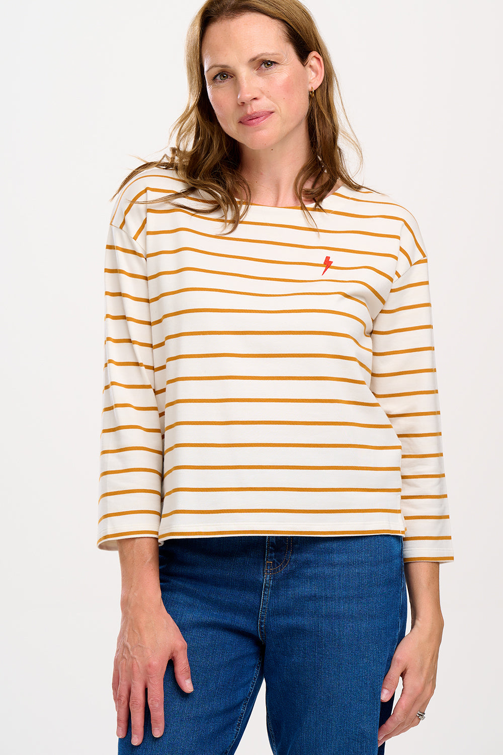 A female model wearing an off-white and tan striped Billie Breton top with a pink embroidered lightning flash on the chest, paired with blue jeans.